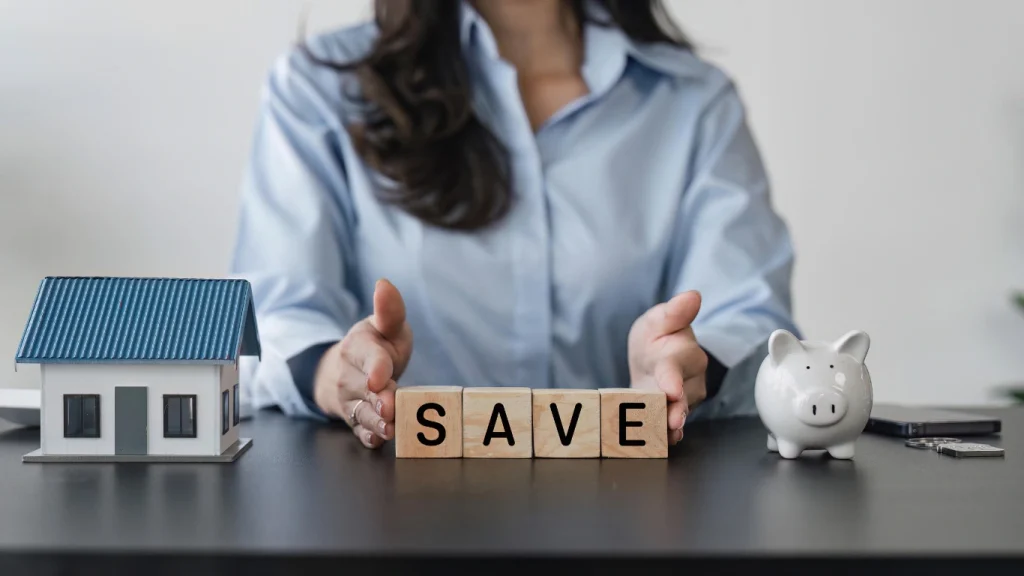 Woman protecting model home and piggy bank with “SAVE” blocks — representing life insurance savings and financial planning.